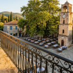 View from a traditional stone balcony in Greece overlooking a village square with a plane tree and clock tower.